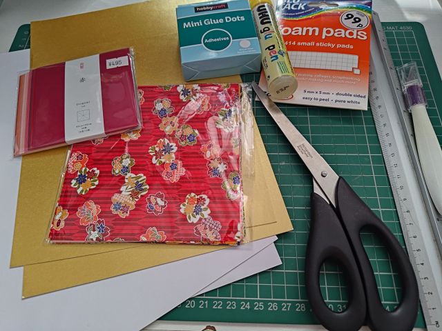 An assortment of craft materials including colourful paper, scissors, adhesive glue dots, foam pads, and a glue stick arranged on a green cutting mat.