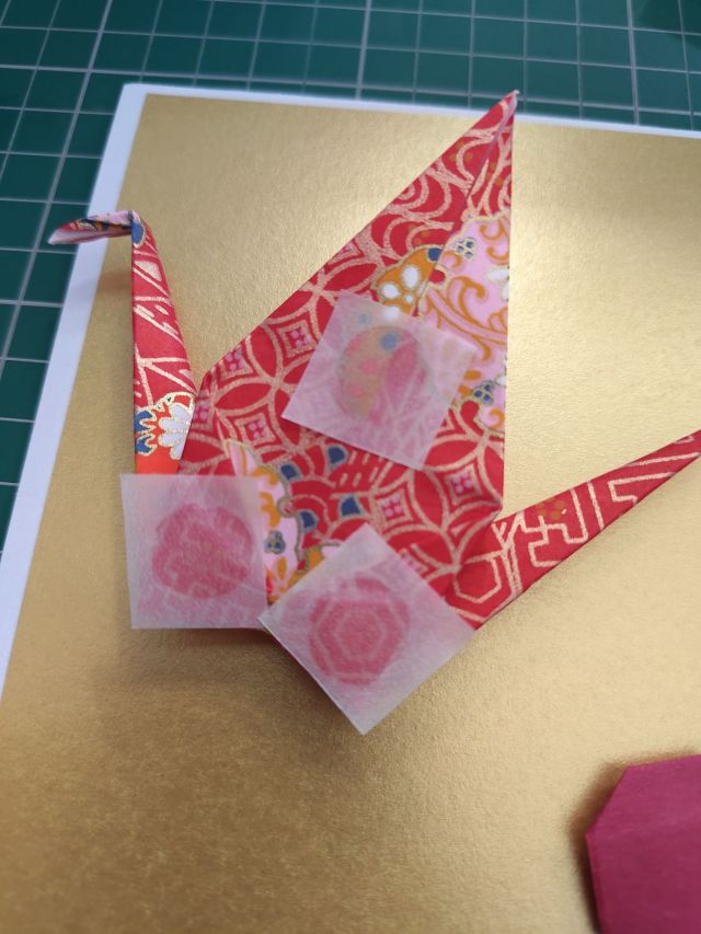 An intricately folded origami crane made from patterned red paper, featuring decorative elements and small translucent squares attached to its body, placed on a gold background.